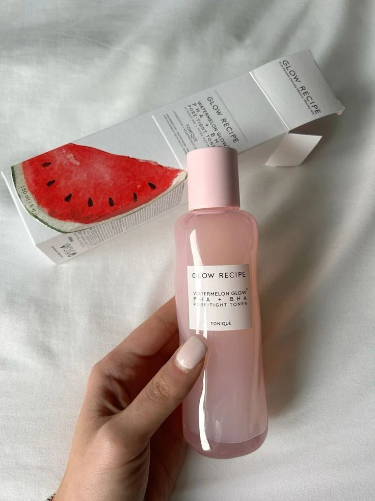 A hand holds a bottle of Glow Recipe Watermelon Glow Skin Toner next to its white packaging box, which features a watermelon illustration, set against a soft white fabric background.