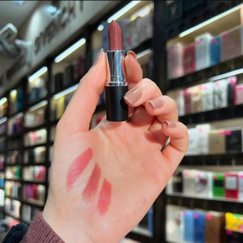 Hand holding a red lipstick with swatches on a blurred store shelf background