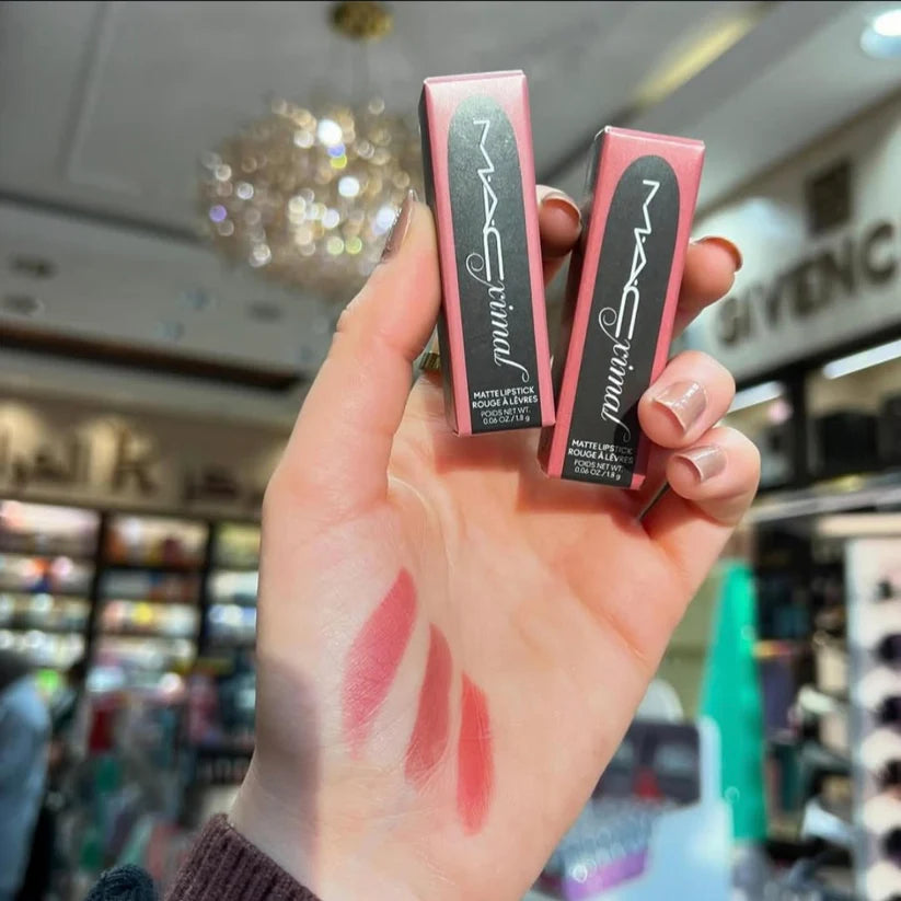 Two MAC Extreme Lipstick boxes held by a hand with lipstick swatches, blurred store interior in the background